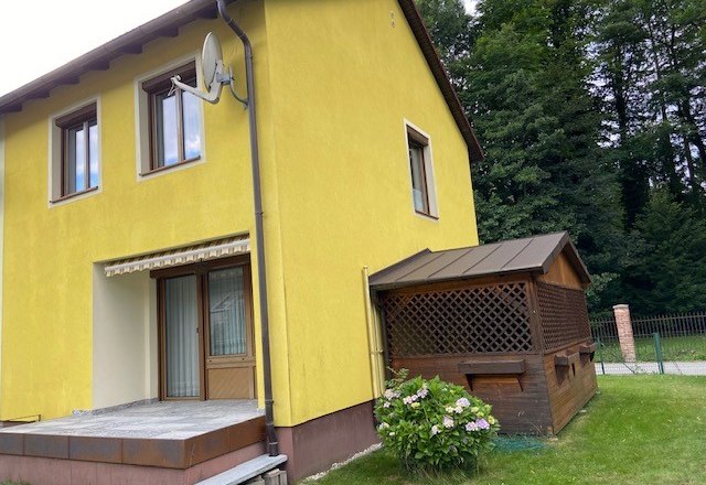 Gelbes Ferienhaus mit Terrasse und kleinem Anbau, umgeben von B&auml;umen.