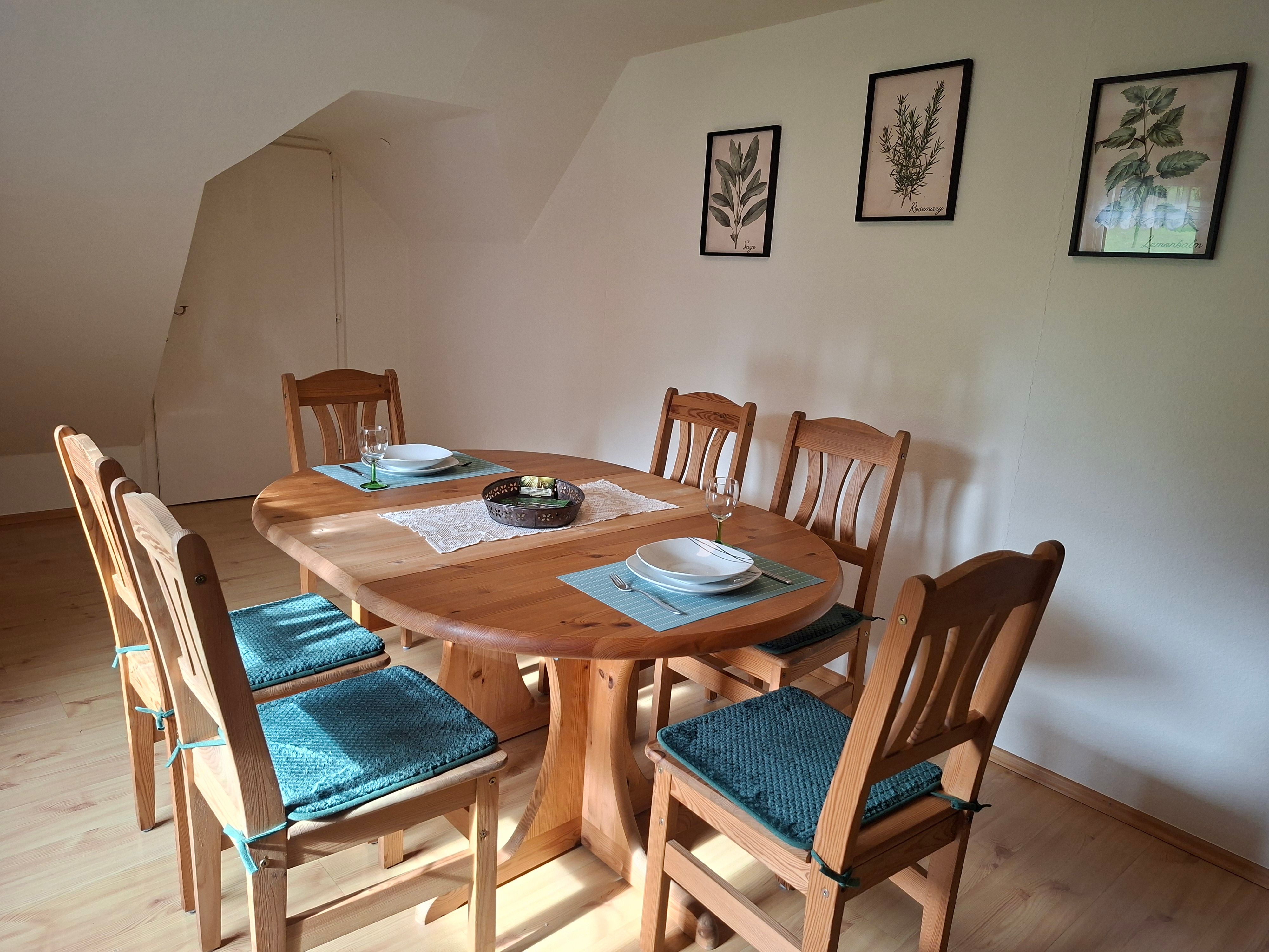 Essbereich mit Holztisch und sechs Stühlen, dekoriert mit blauen Sitzkissen. An der Wand hängen drei botanische Bilder.