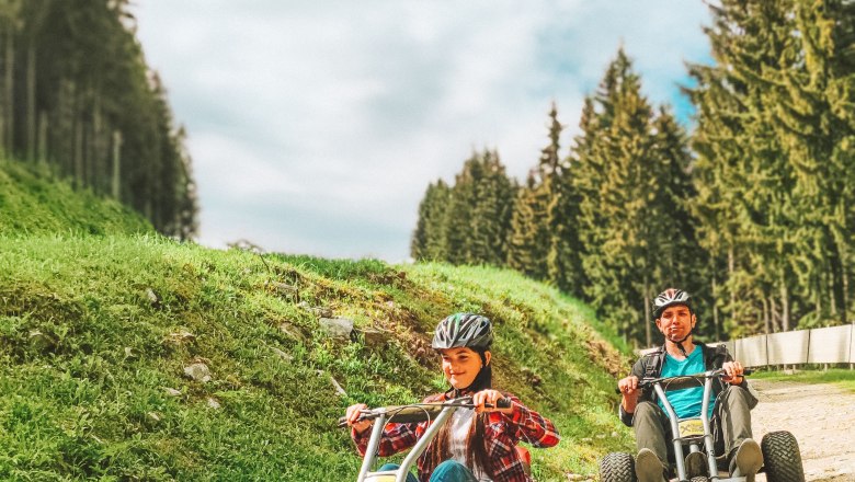 Zwei Personen fahren Mountaincarts auf einem Schotterweg, umgeben von Bäumen und blauem Himmel.
