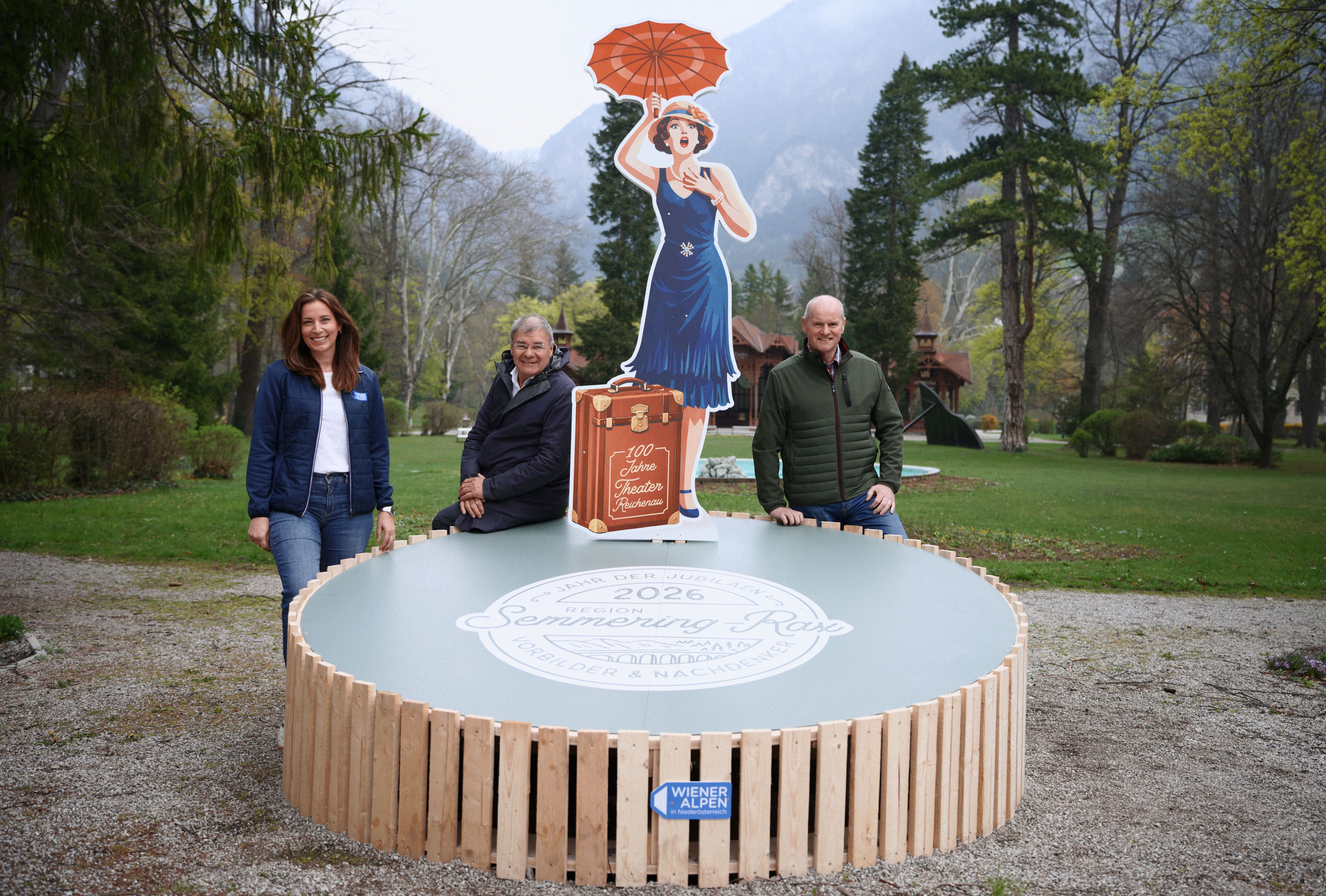 Drei Personen stehen rund um eine überlebensgroße, zweidimensionale „Jubiläumsfigur" - eine schick gekleidete Dame mit Hut, Schirm und Koffer - im Kurpark Reichenau