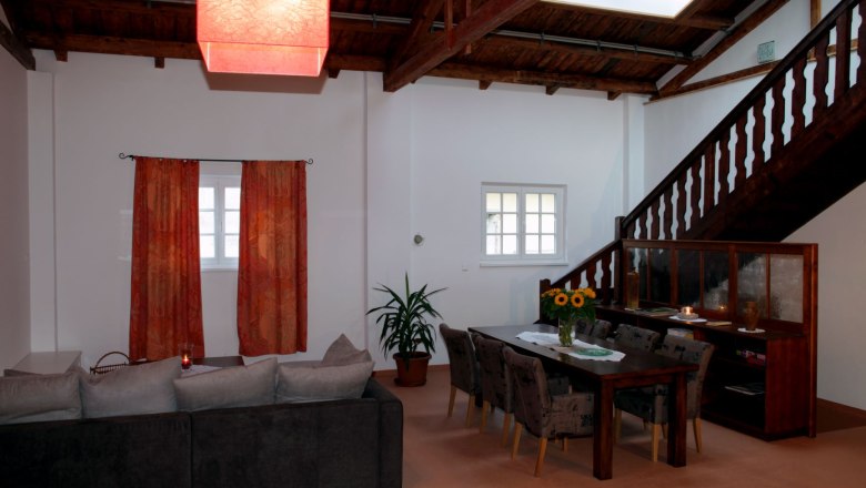 Wohn- und Essbereich in einem Loft mit Holzbalkendecke, rotem Lampenschirm, grauem Sofa, Esstisch mit Stühlen und Treppe.