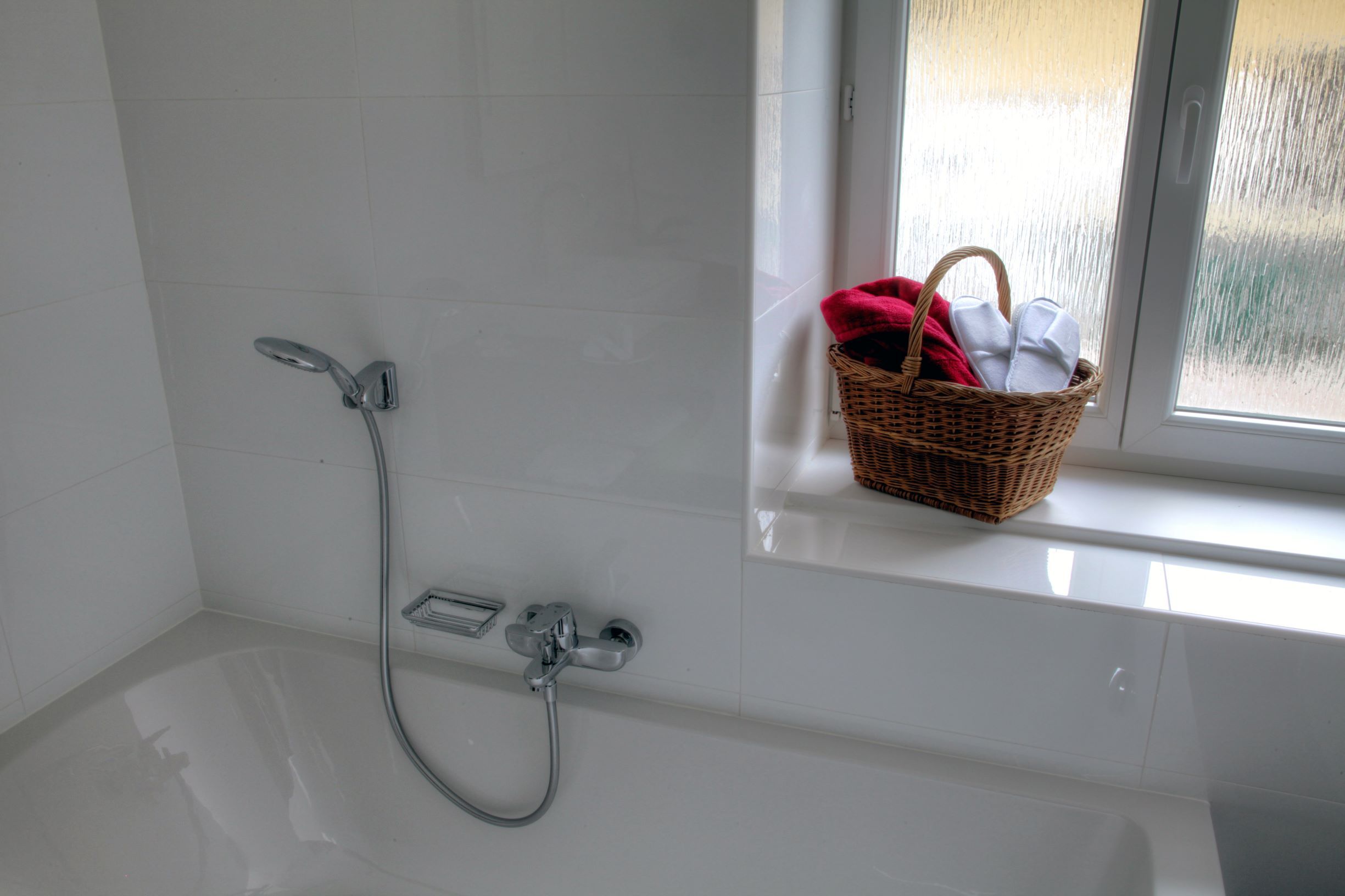 Weiße Badewanne mit Duschkopf, daneben ein Fensterbrett mit einem Korb voller Handtücher.