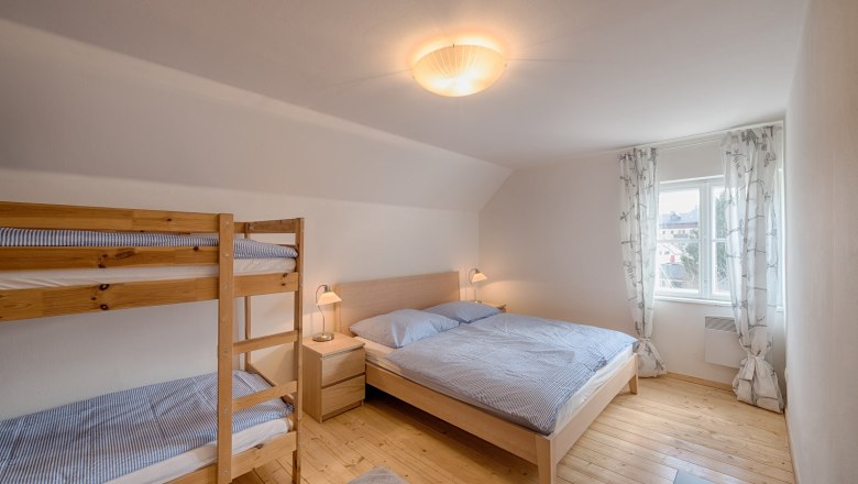 Helles Schlafzimmer mit Doppelbett und Etagenbett, Holzboden und Fenster mit Vorhängen.