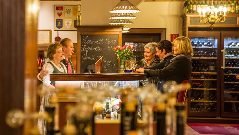 Menschen in einer gem&uuml;tlichen Gaststube mit Weinflaschen im Hintergrund.