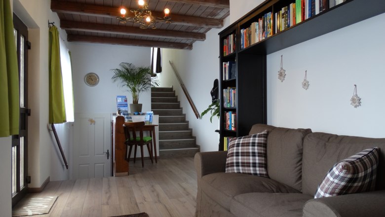 Wohnzimmer mit braunem Sofa, Bücherregal und Treppe im Hintergrund.