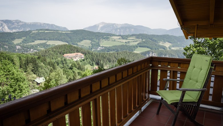 Blick von einem Balkon auf eine Berglandschaft mit Wäldern und einem Gebäude in der Ferne.