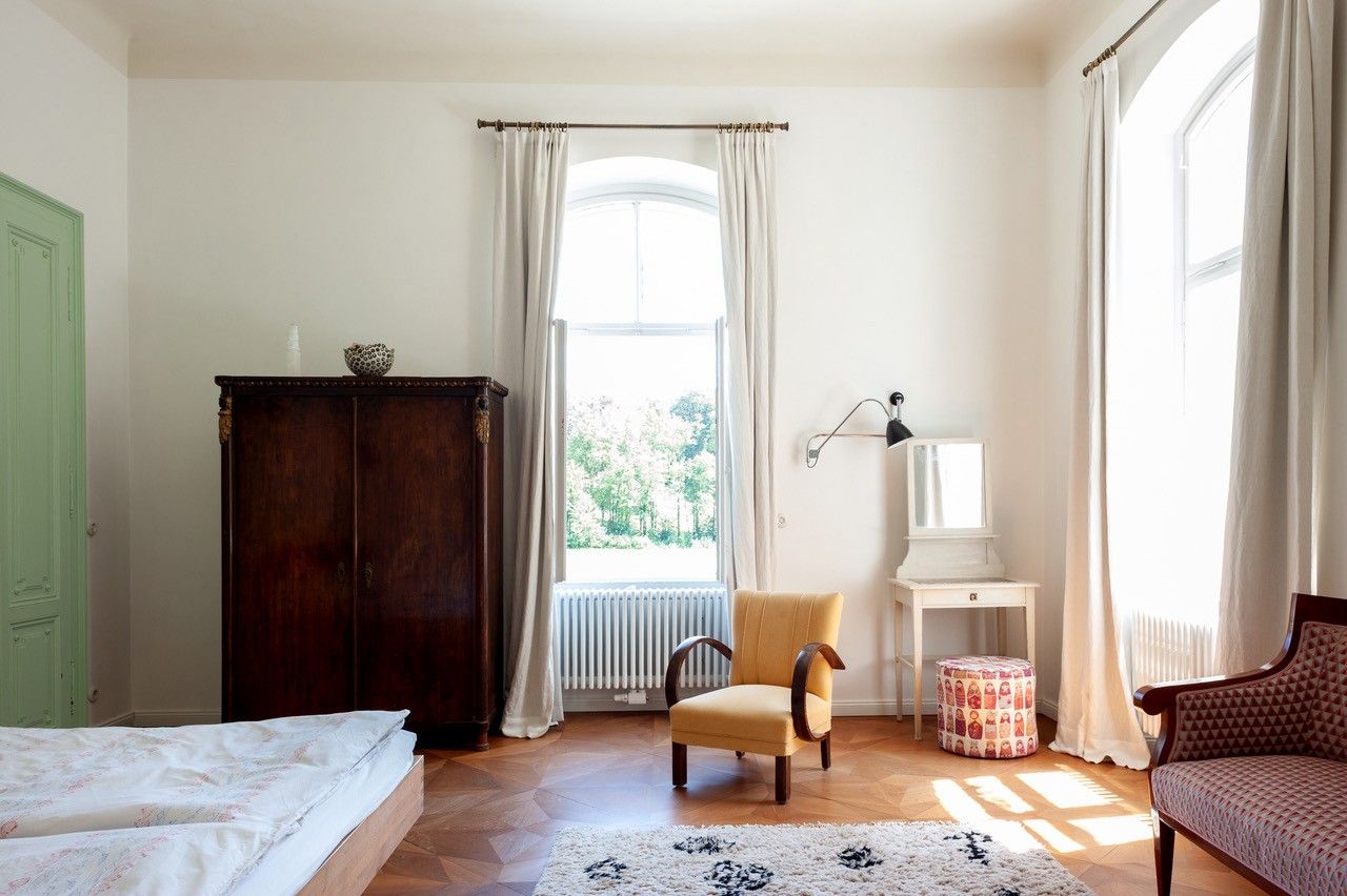 Ein helles Schlafzimmer mit einem großen Fenster, einem antiken Schrank, einem gelben Sessel und einem kleinen Schminktisch.