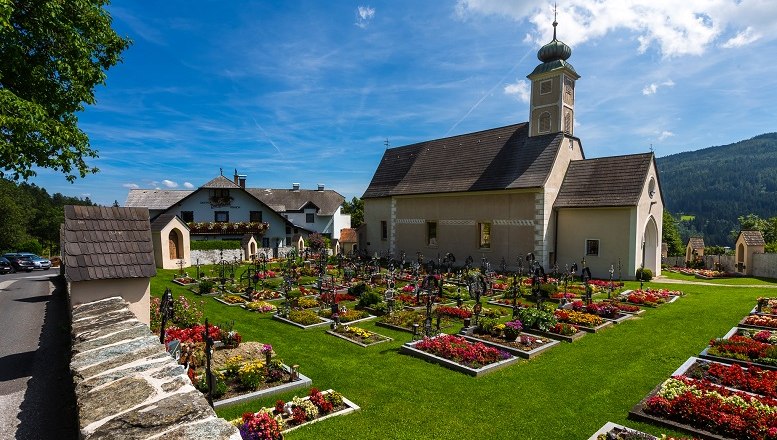 Parish church of St. Peter am Neuwald with a well-tended cemetery and flower beds in bloom, surrounded by a green landscape and blue sky.