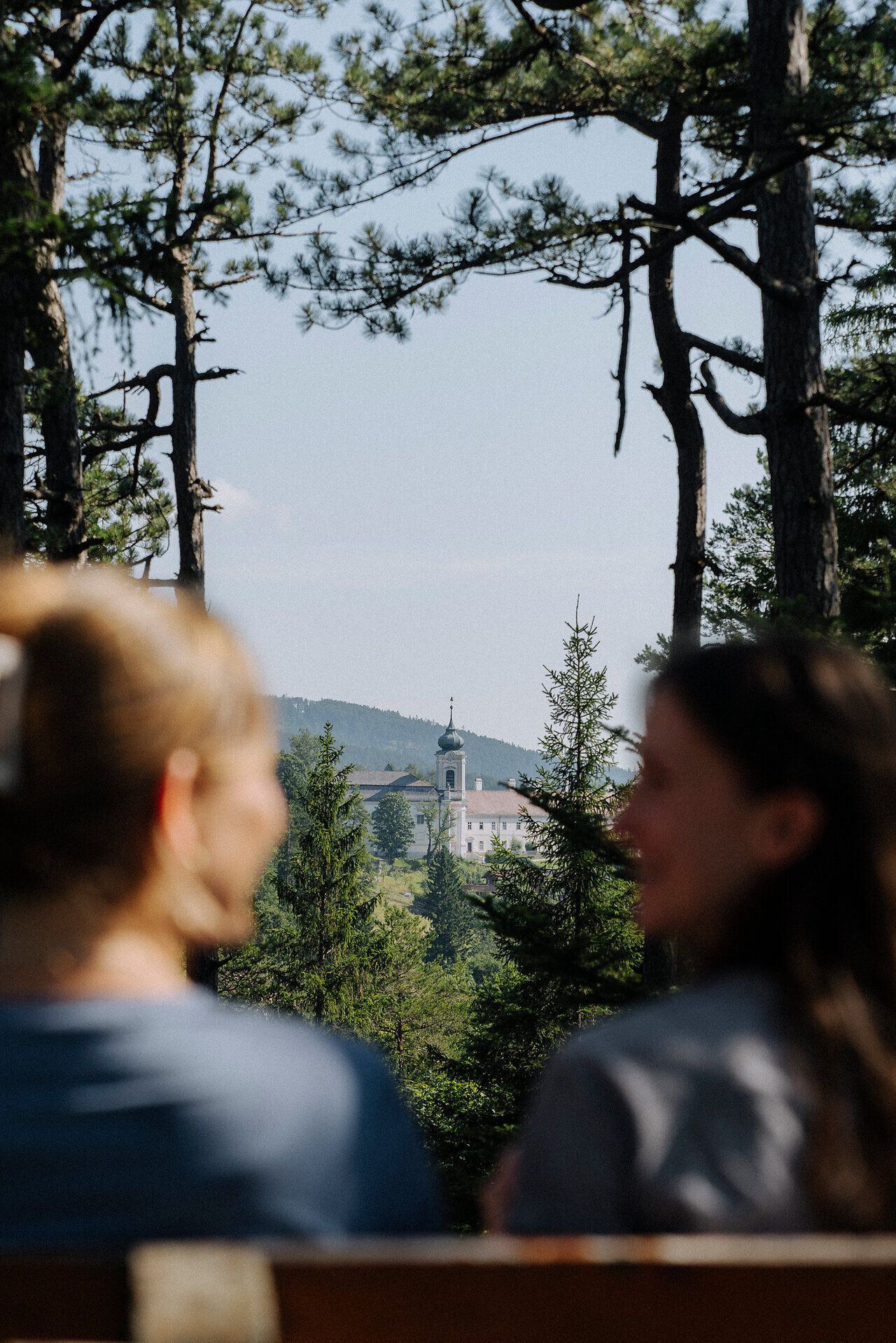 Im Vordergrund sitzen zwei Frauen, auf einer Bank und unterhalten sich. Dahinter sieht man im Fokus den Blick auf Gutenstein in der Ferne.