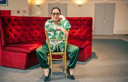 Frau mit Brille sitzt lässig auf einem Stuhl vor einer roten Samtbank in einem modernen Innenraum.