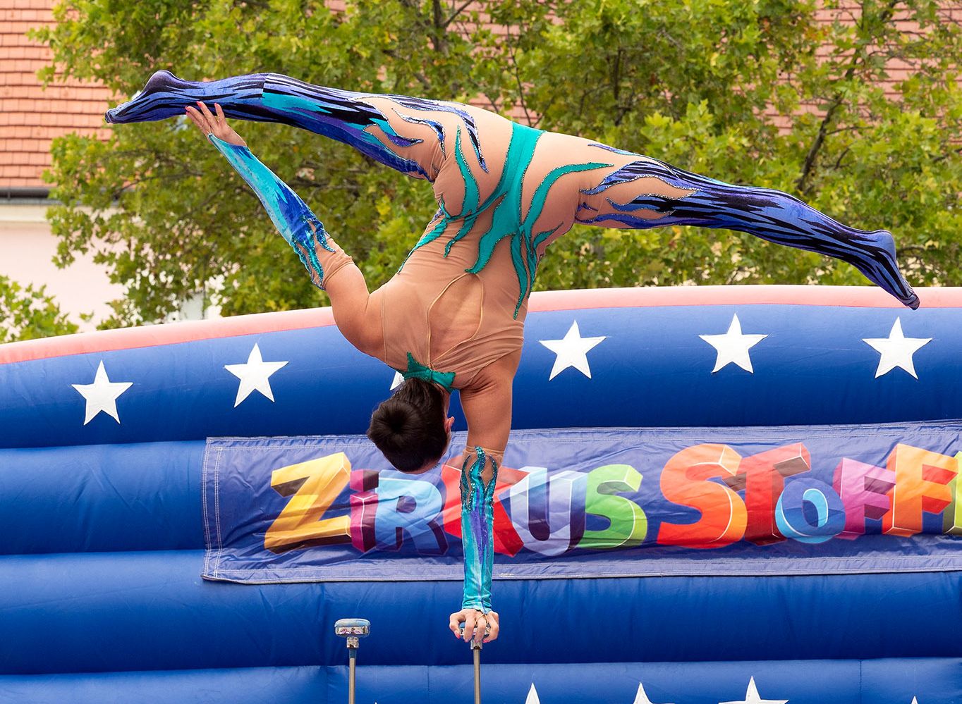 Akrobat turnt in einem blauen Kostüm auf einem Stab. Im Hintergrund ist eine blaue Hüpfburg. 