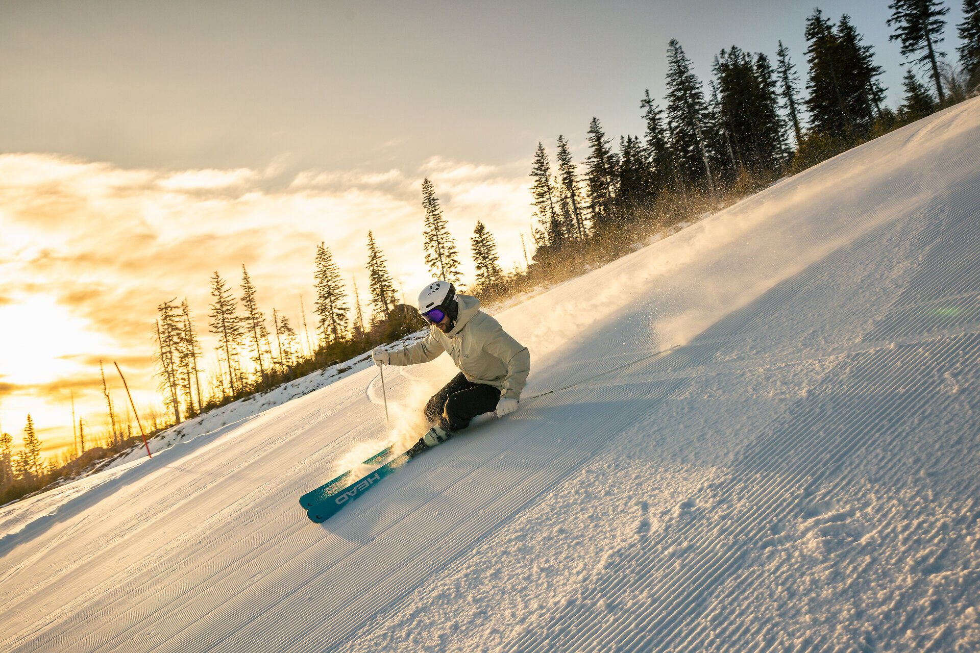 Skifahrende Person im Vordergrund auf einer präparierten Skipiste mit Blick über verschneite Berghänge und Wolken bei tief stehender Sonne.