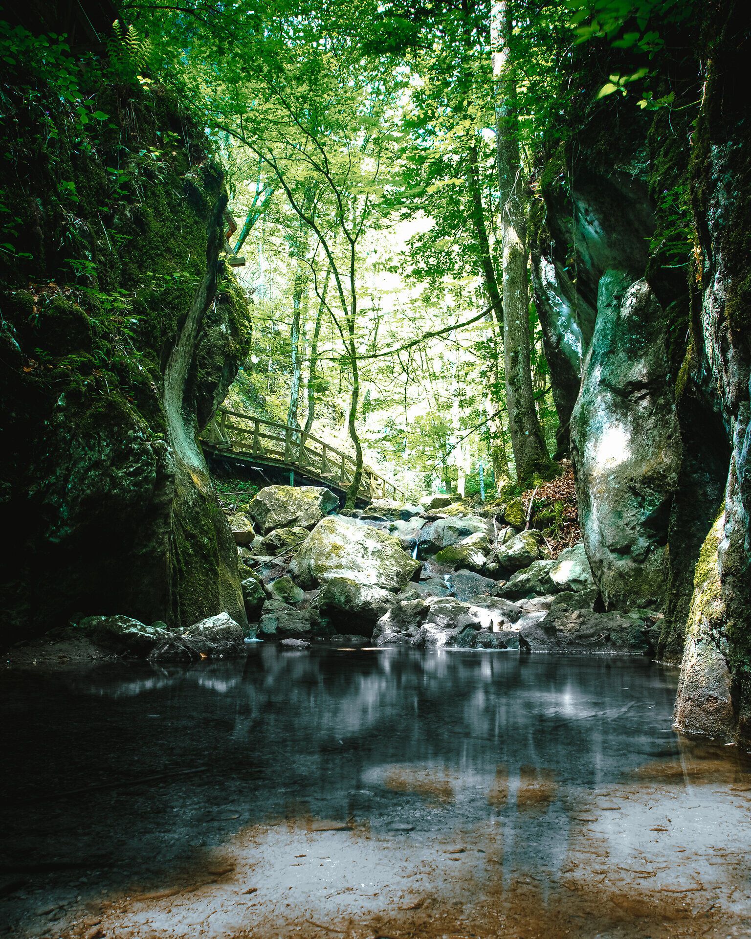 Die sanften Lichtstrahlen durchdringen das dichte Blätterdach und zaubern ein magisches Spiel aus Licht und Schatten auf die glitzernden Wasseroberflächen. Umgeben von majestätischen Felsen und üppigem Grün lädt dieser Ort zum Verweilen und Entspannen ein, während das sanfte Plätschern des Wassers eine beruhigende Melodie spielt.
