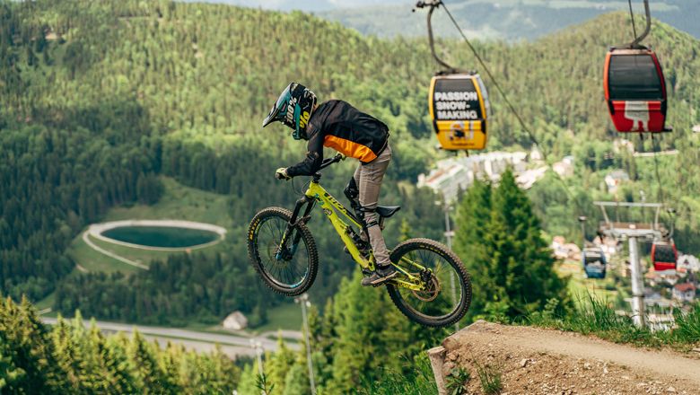 Mountain biker jumping in the Semmering bike park, cable car and forest in the background.