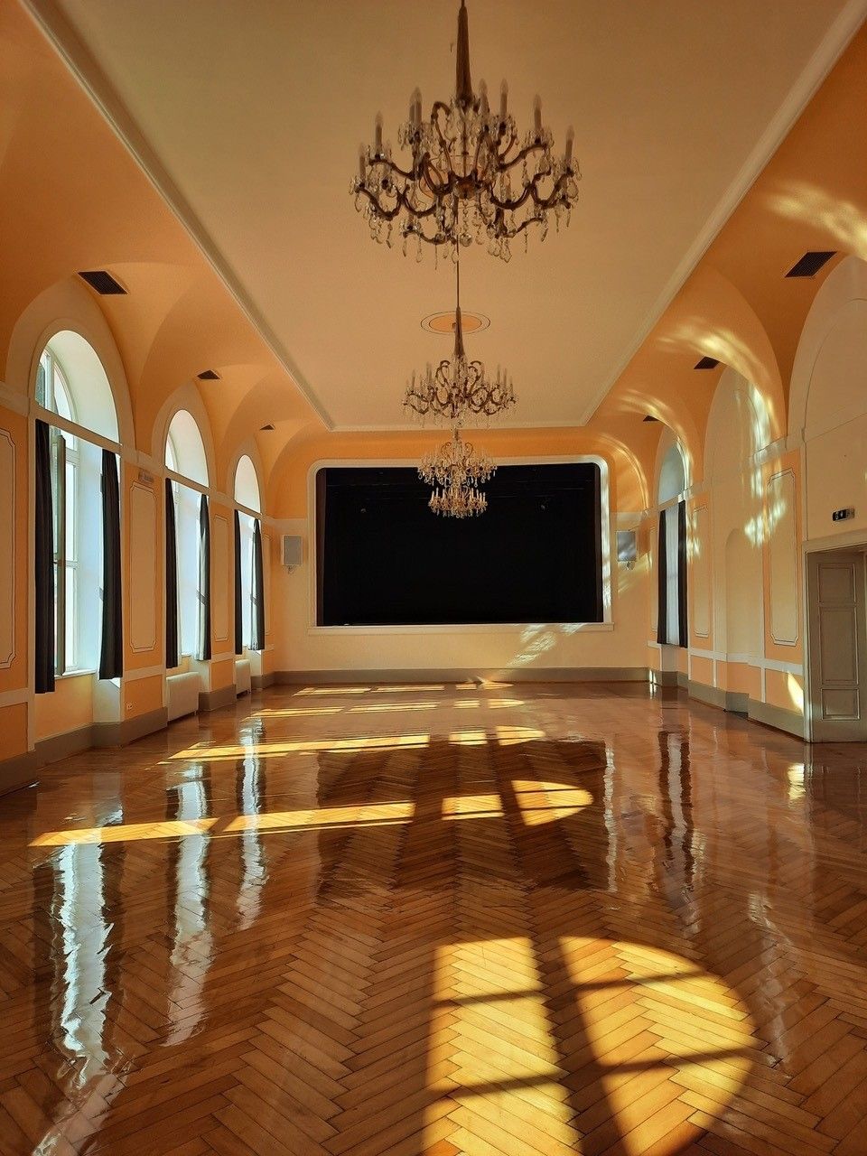 Ein Saal mit Bühne, Parkettboden, Kronleuchtern und großen Fenstern, durch die die Sonne scheint.