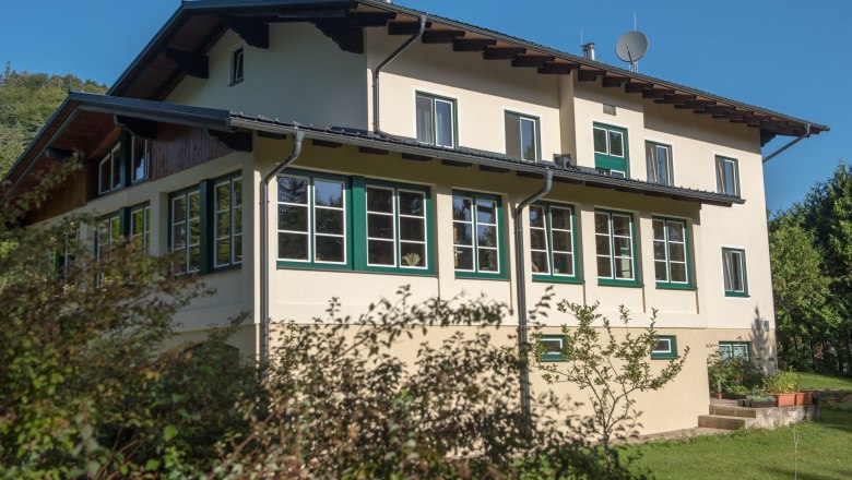 Ein zweist&ouml;ckiges Haus mit vielen Fenstern und einem Satteldach, umgeben von B&auml;umen und Str&auml;uchern, unter einem klaren blauen Himmel.