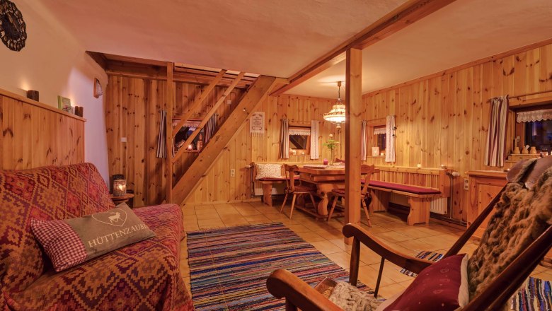 Gemütliches Wohnzimmer mit Holzwänden, Sofa, Schaukelstuhl und Esstisch. Rustikales Ambiente mit Teppich und warmem Licht.