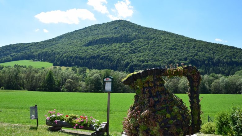 Grüne Landschaft mit einem bewaldeten Hügel im Hintergrund und einer bepflanzten Skulptur im Vordergrund.