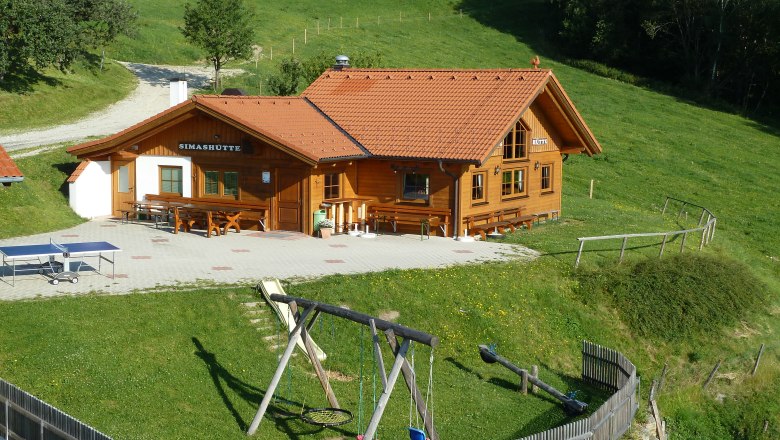 Eine Berghütte namens Simashütte mit rotem Dach, umgeben von grünen Wiesen und einem Spielplatz mit Schaukel.