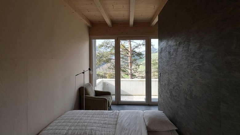 Minimalistisches Schlafzimmer mit Bett, Sessel und Fenster mit Blick auf Bäume.