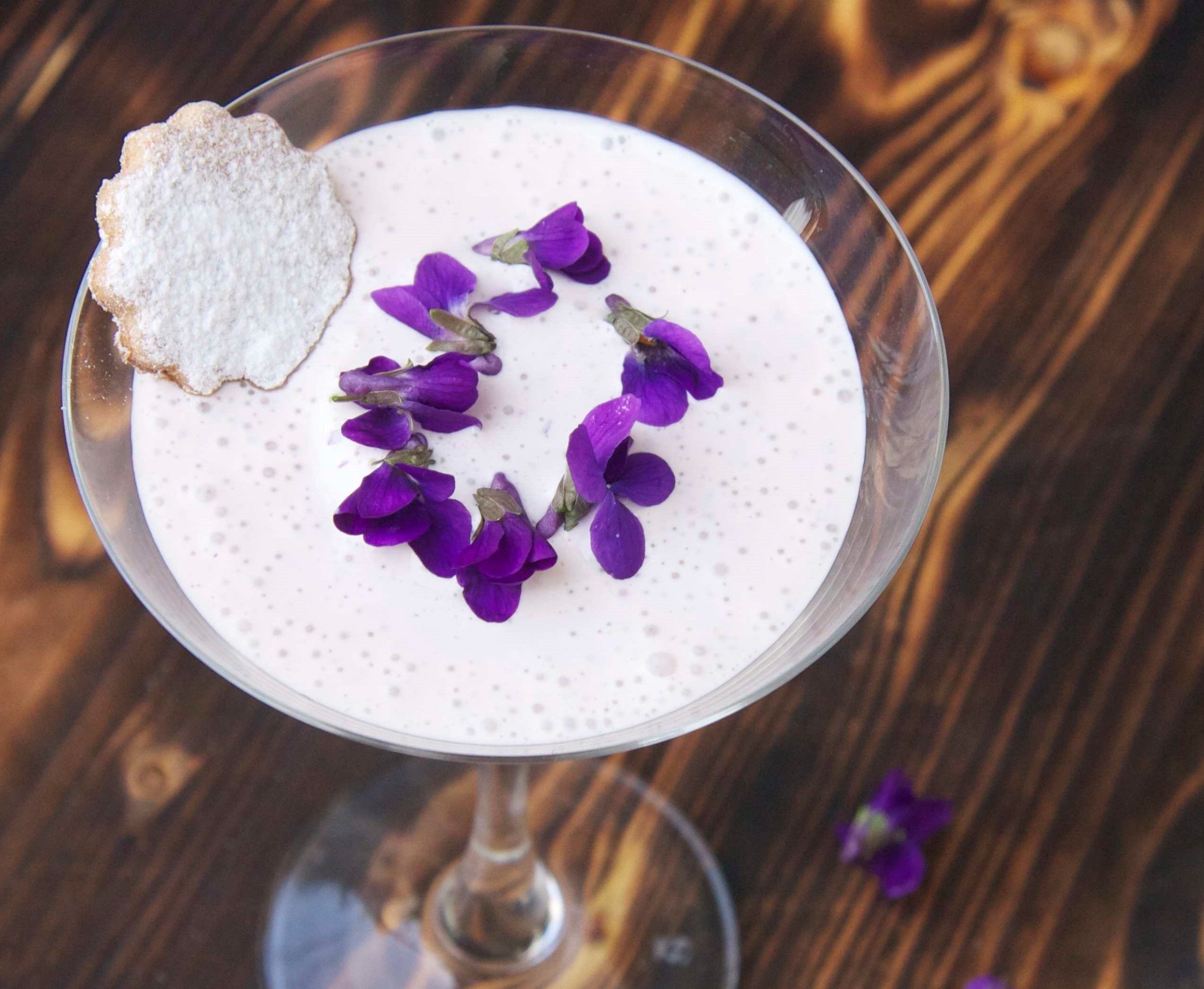 Glas mit Veilchen-Creme, Veilchenblüten und Mürbteigkeks auf rustikalem Holztisch.