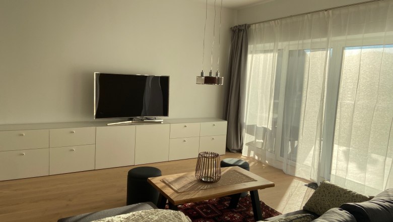 Moderner Wohnbereich mit grauem Sofa, Holztisch, Teppich, Fernseher und großen Fenstern mit Vorhängen.