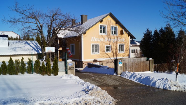 Hausansicht im Winter, © Reep Ein gelbes Haus mit der Aufschrift 'Haus Bergblick' im Schnee, umgeben von Bäumen und einem blauen Himmel.