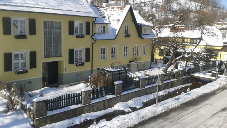 Gelbes Haus im Winter mit Schnee bedeckt, umgeben von einem Zaun und Bäumen.