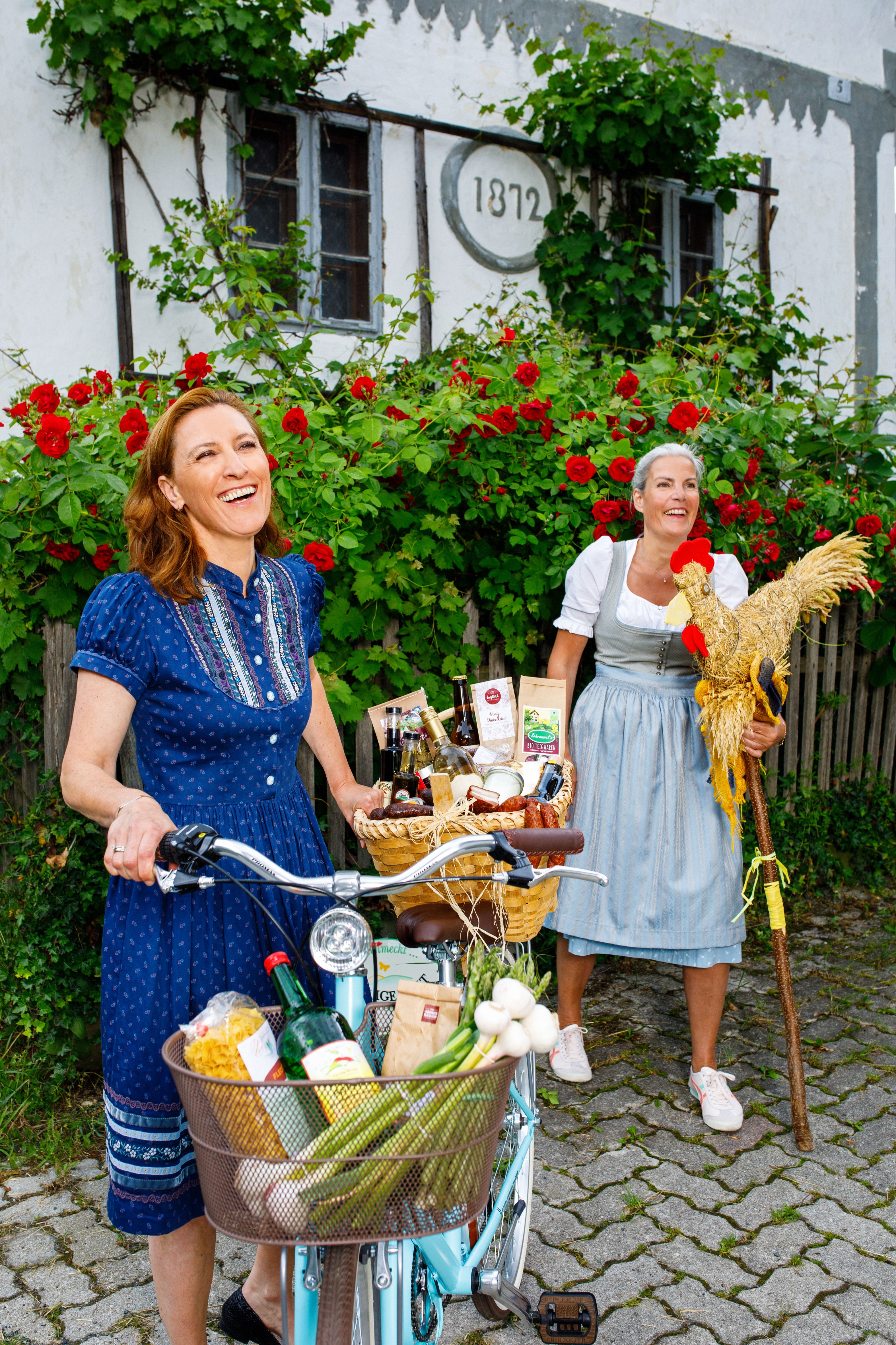 Zwei Frauen in Tracht gekleidet, eine hält ein Fahrrad, die andere einen Schnidahahn aus Stroh.