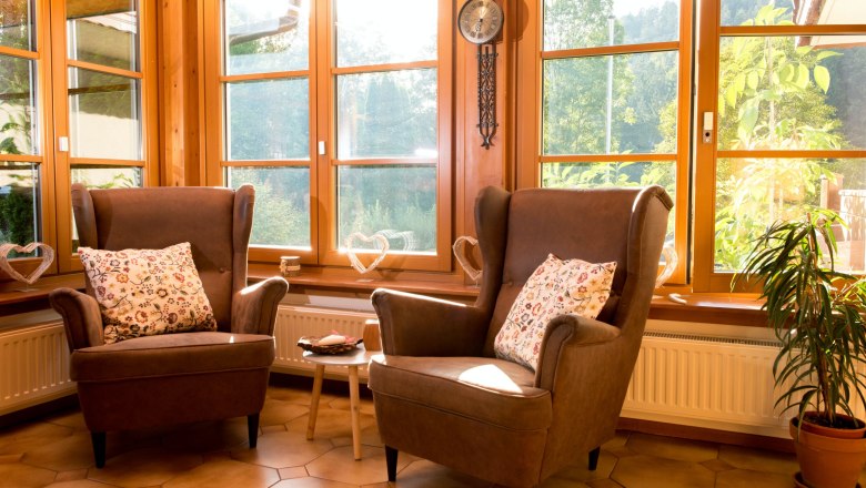 Zwei braune Sessel mit gemusterten Kissen vor gro&szlig;en Fenstern in einem sonnigen Raum.