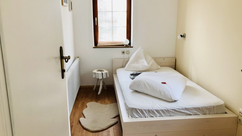 A small, cozy bedroom with a single bed, wooden floor and a small window with blinds.