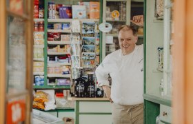 Ein Mann in Kochkleidung steht lächelnd in einem alten Laden mit Regalen voller Produkte.