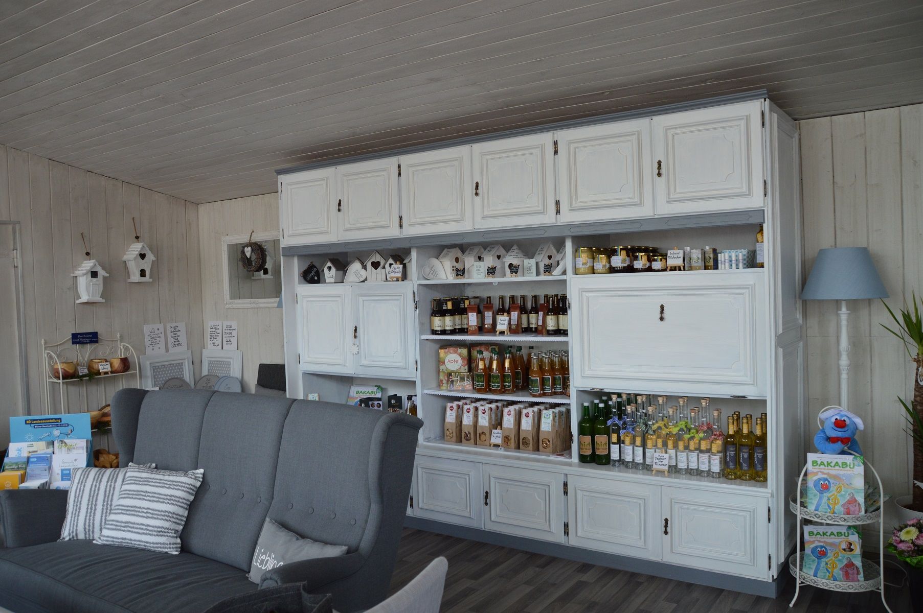 Innenansicht eines gemütlichen Ladens mit weißen Regalen, gefüllt mit Getränken und Produkten. Ein graues Sofa steht im Vordergrund.