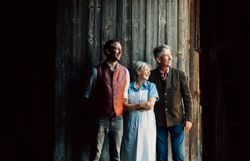 Drei Personen stehen lächelnd vor einer Holzwand und schauen nach rechts.