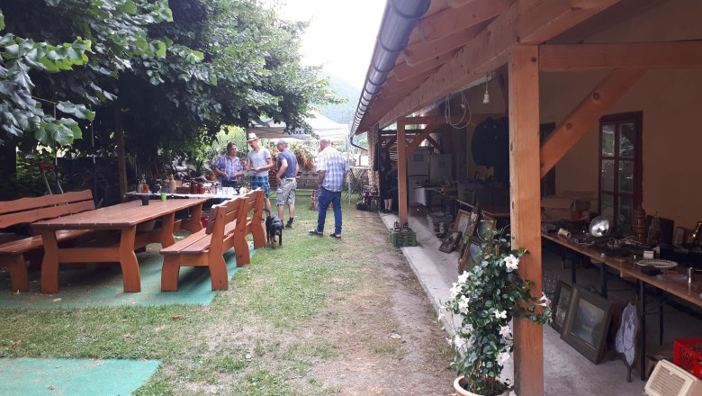 Ein Gastgarten mit Holztischen und Bänken, Menschen im Gespräch, ein Hund, und ein überdachter Bereich mit Flohmarkt.