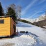 Kleines Holzhaus auf schneebedecktem Hügel mit Berglandschaft im Hintergrund.