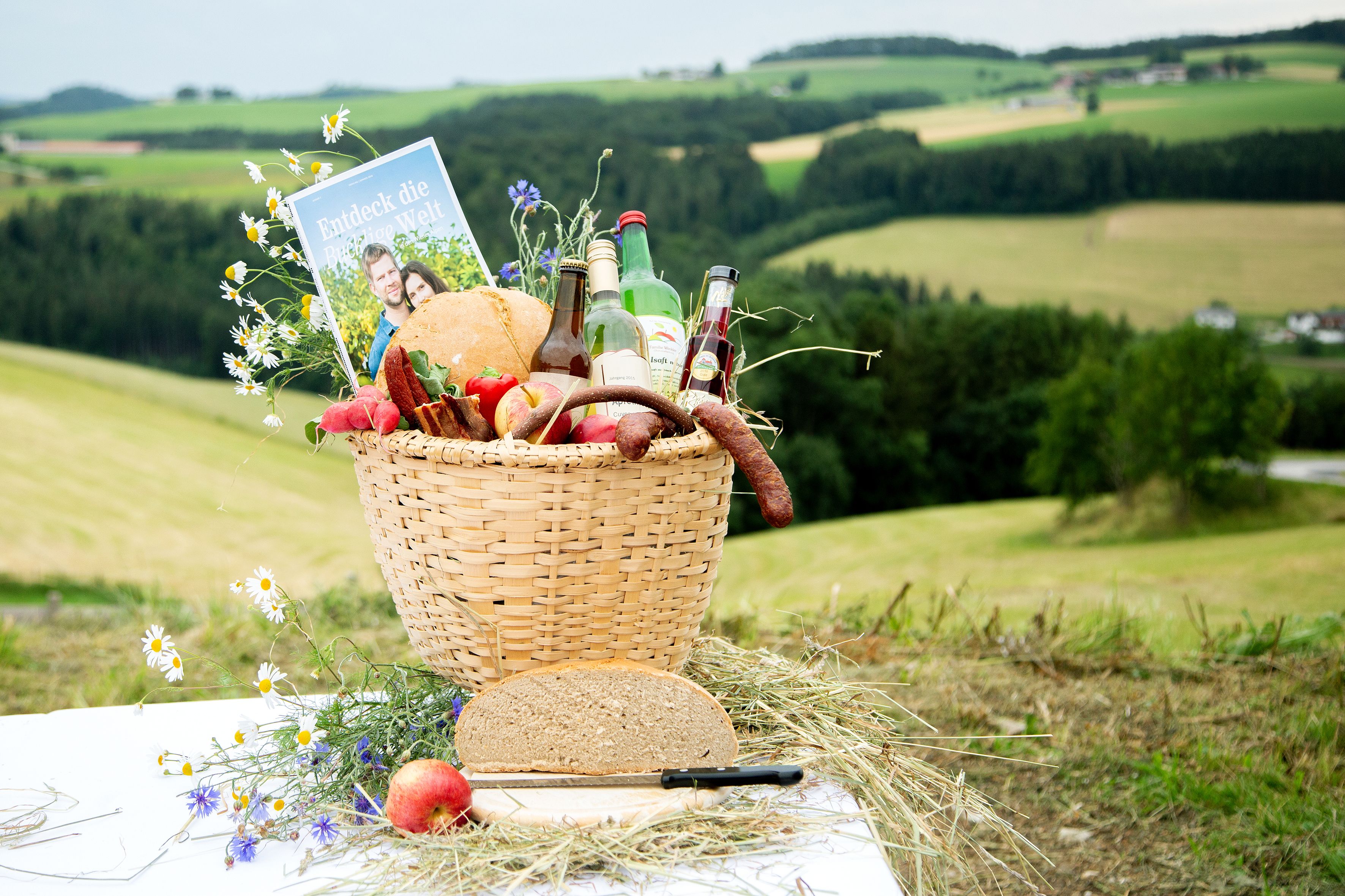 Ein Bucklkorb mit regionalen Lebensmitteln, davor Brot und ein Apfel, im Hintergrund grüne Landschaft.