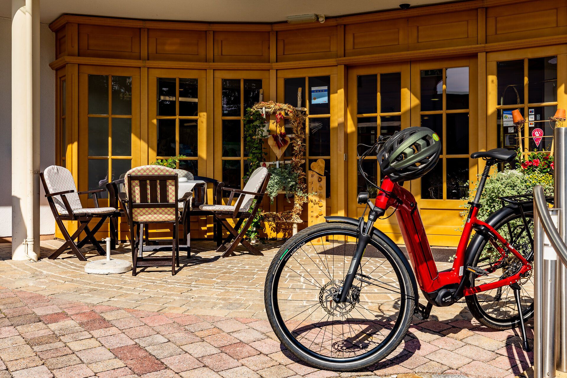 Ein rotes Fahrrad parkt mit Helm auf der Lenkstange auf der Terrasse vom Vitalzeit Hotel Weber. Vor der hellen Holzfenster-Front steht ein Tisch und Stühle mit Polstern. 