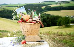 Ein Bucklkorb mit regionalen Lebensmitteln, davor Brot und ein Apfel, im Hintergrund grüne Landschaft.