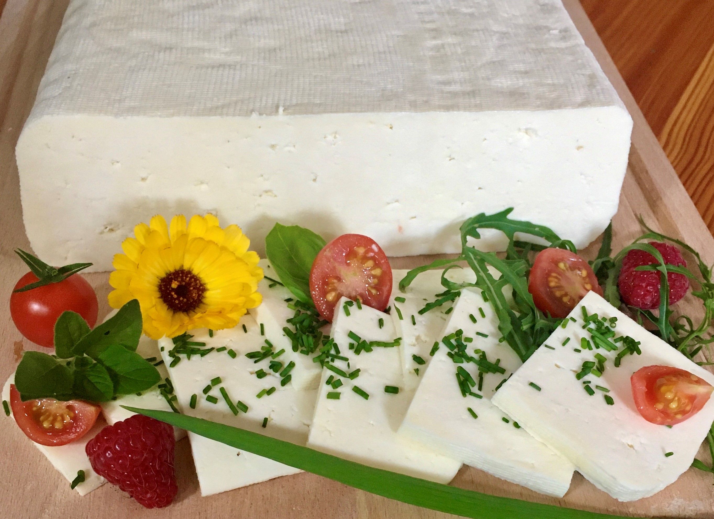 Ein Block Schafkäse in Scheiben geschnittenen, garniert mit Tomaten, Himbeeren, Rucola und einer gelben Blume auf einem Holzbrett.