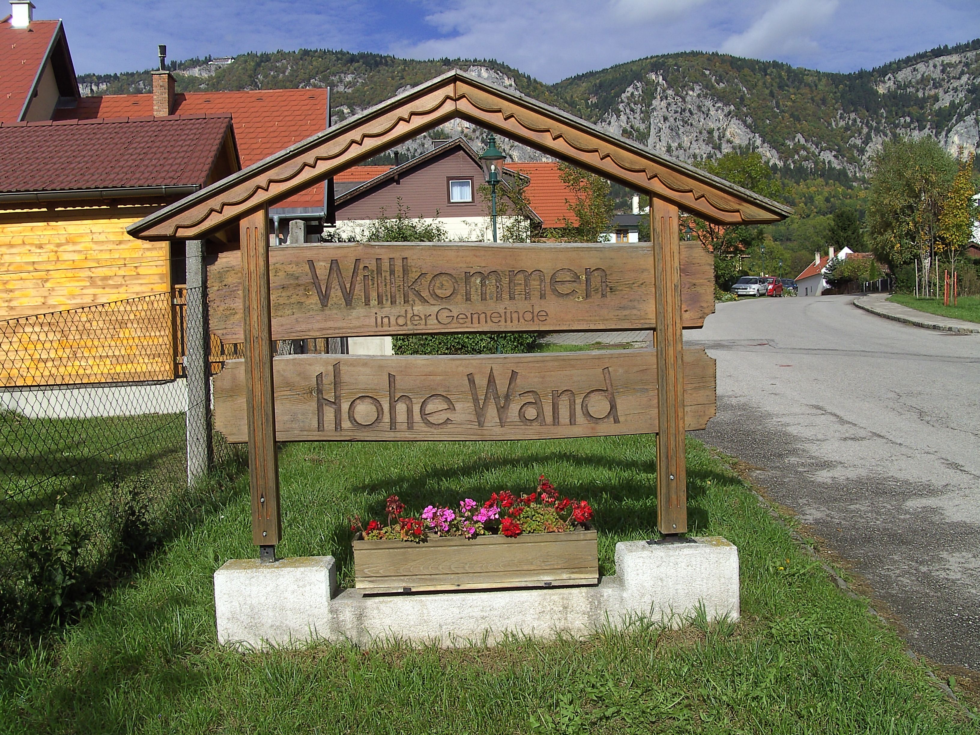 Holzschild mit der Aufschrift 'Willkommen in der Gemeinde Hohe Wand' vor einer Berglandschaft.