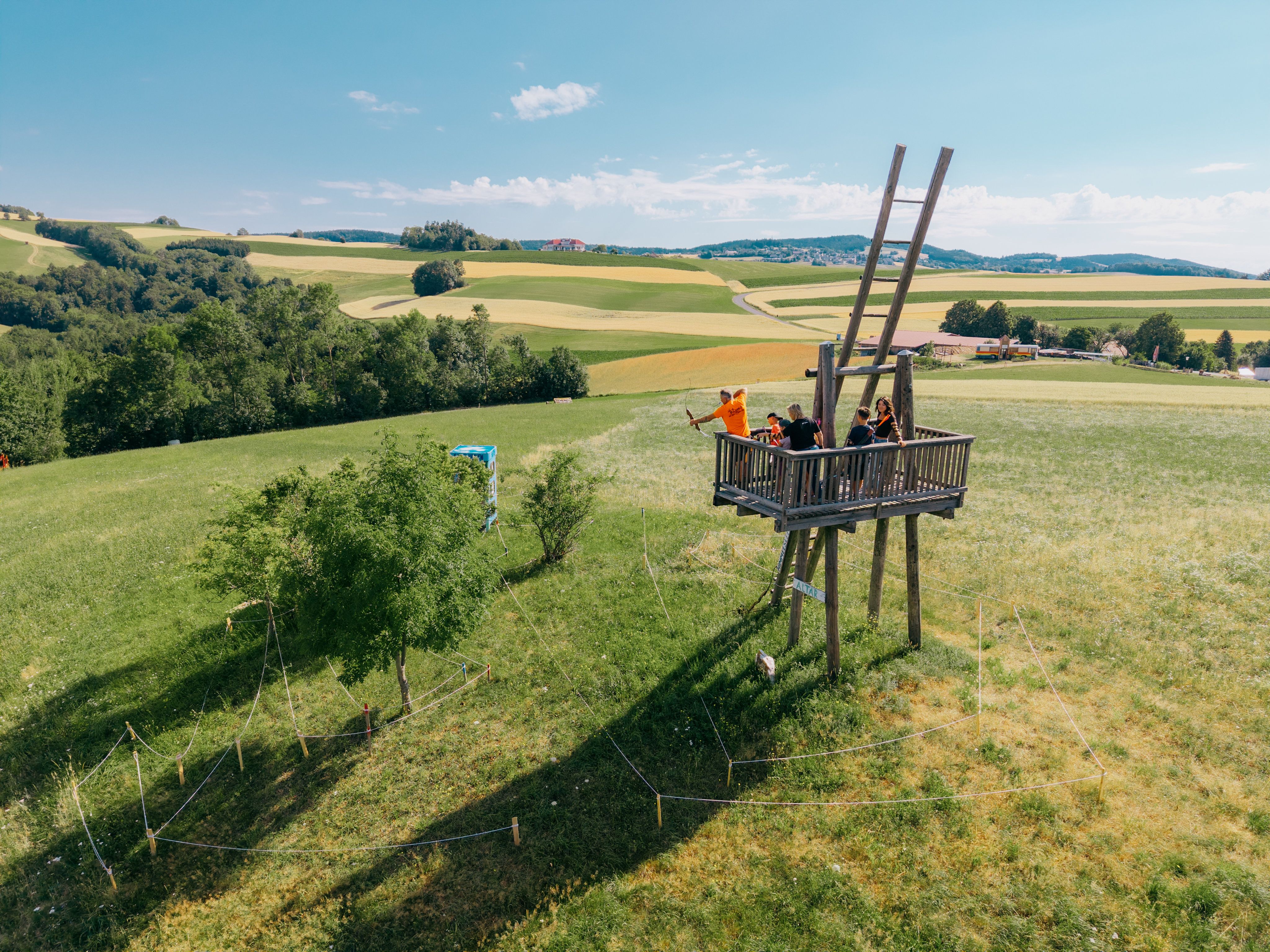 Menschen auf einem Holzturm beim Bogenschießen in einer ländlichen Landschaft.