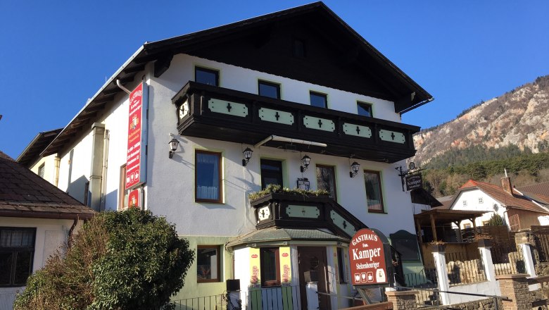 Ein traditionelles Gasthaus mit Holzverzierungen und einem Schild "Gasthaus Fam. Kamper" vor einem klaren blauen Himmel.