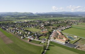 Luftaufnahme eines Dorfes mit Feldern und Hügeln im Hintergrund.