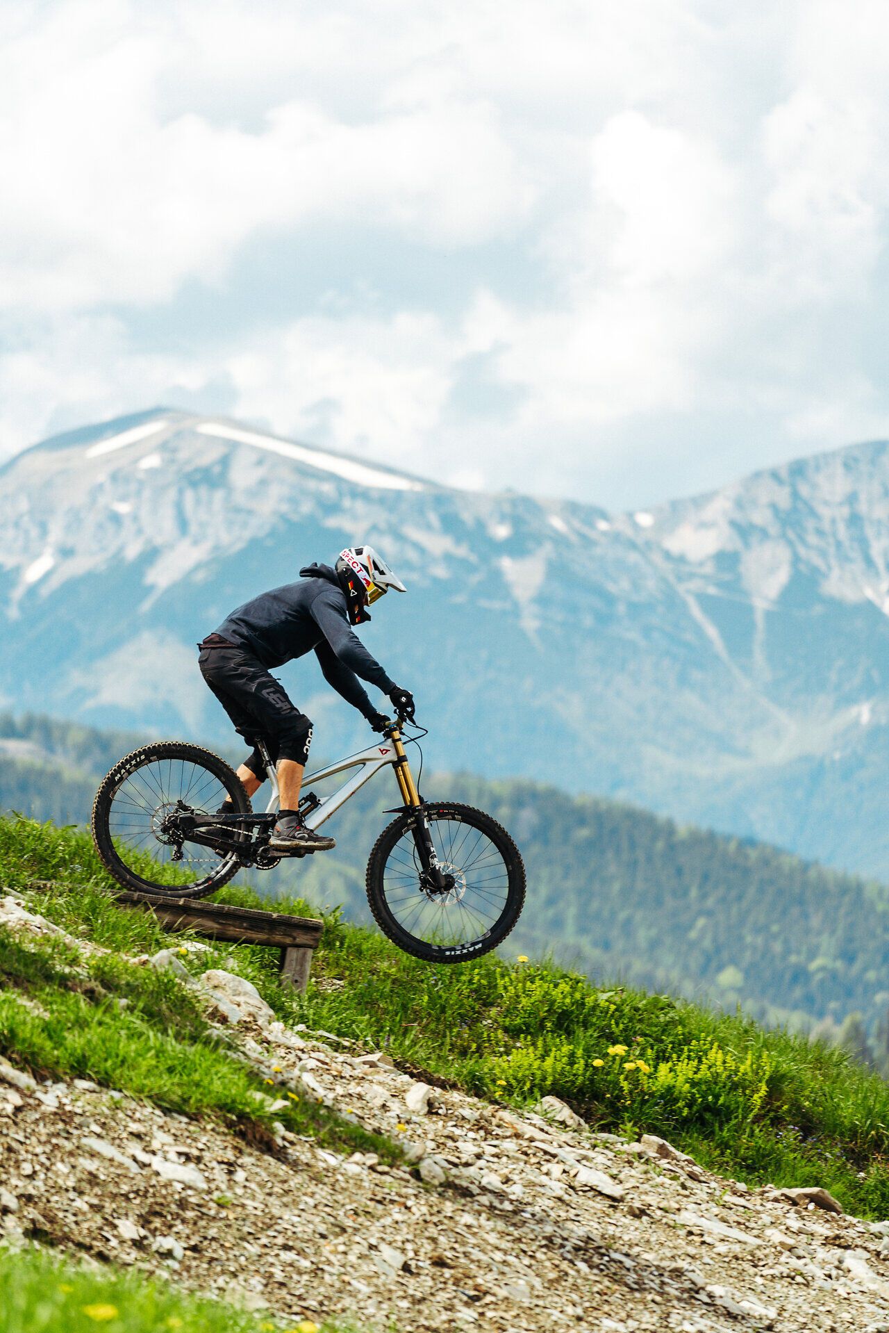 Mountainbiker fährt auf einem schmalen Trail durch alpine Landschaft mit Bergen im Hintergrund.