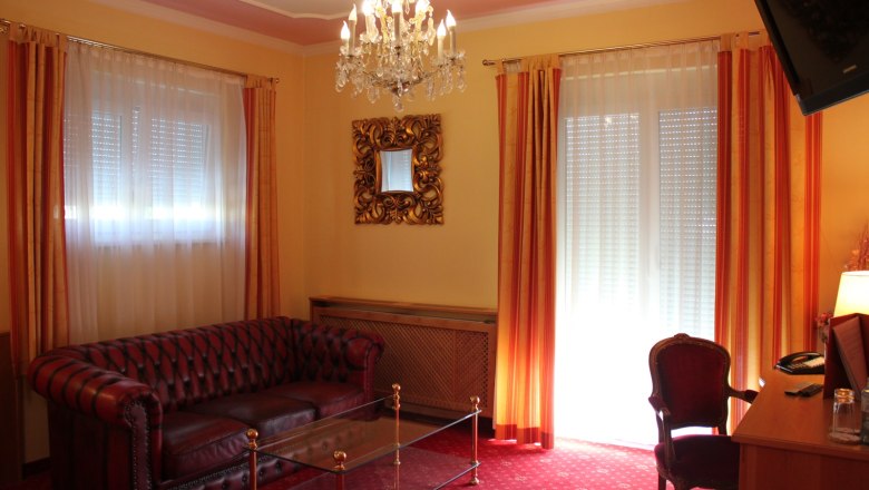 Elegantes Wohnzimmer mit Ledersofa, Glastisch, Kronleuchter und roten Vorh&auml;ngen.