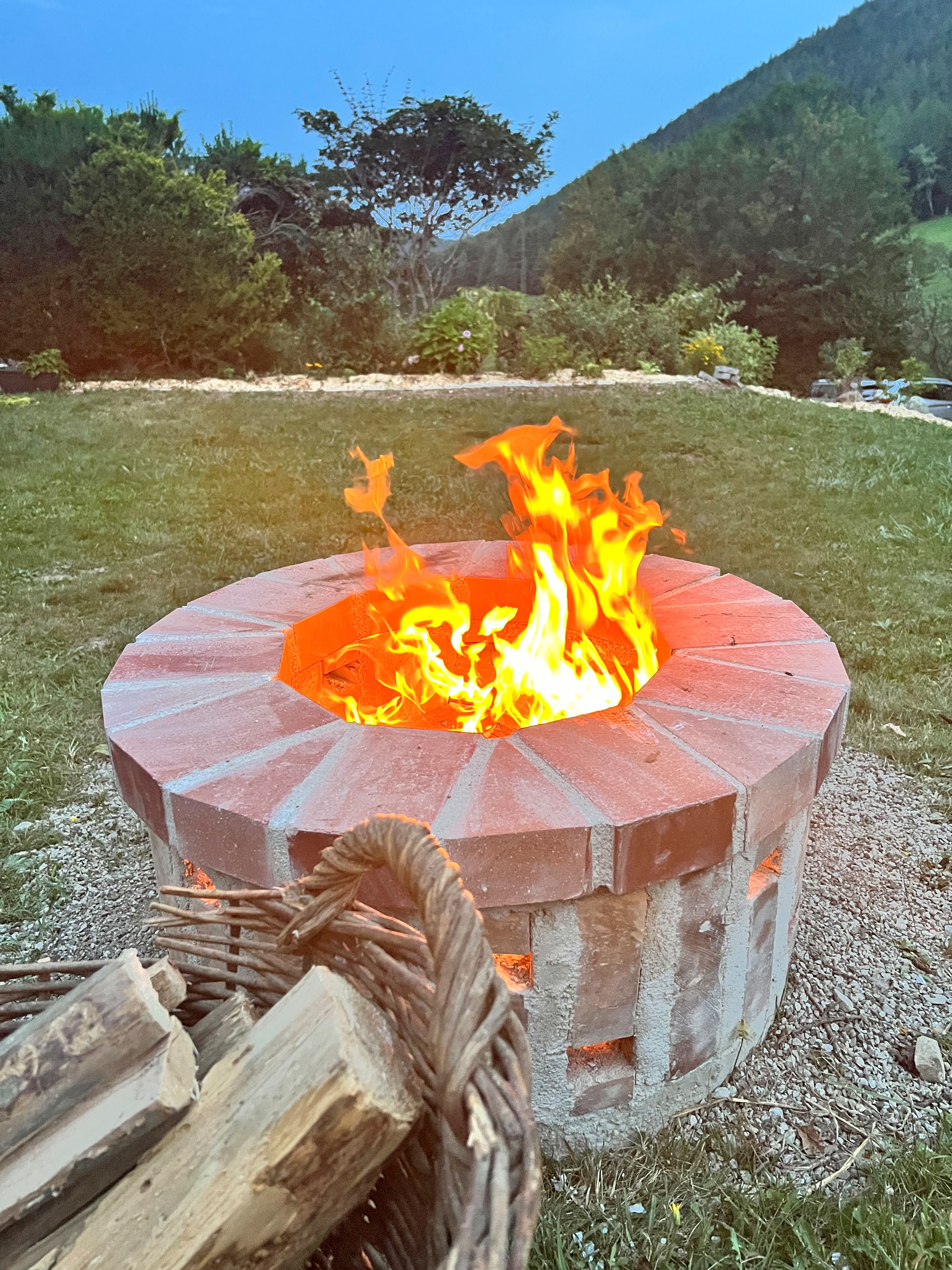 A brick fire pit with a blazing fire in the garden, with a basket of firewood in front of it; in the background, a meadow, trees, and a wooded hillside.