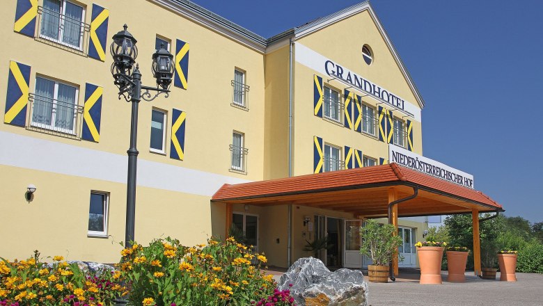 Außenansicht eines gelben Hotels mit blauen Fensterläden und einem Vordach. Vor dem Gebäude blühen gelbe und pinke Blumen.