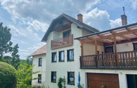 Ein zweistöckiges Haus mit Balkon und Terrasse, umgeben von Garten und Bäumen, unter einem bewölkten Himmel.