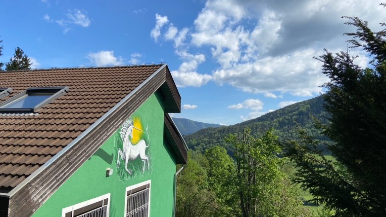 Grünes Haus mit Einhornbild an der Wand, umgeben von Bäumen und Bergen unter blauem Himmel.